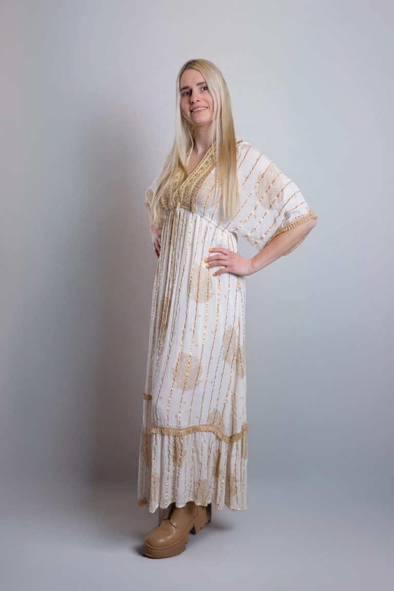 Frau mit langen blonden Haaren trägt ein weißes, gemustertes Maxikleid und Schuhwerk.