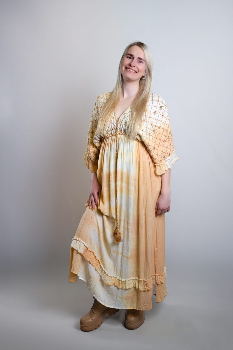 Frau mit langen blonden Haaren in einem langen, orangefarbenen Kleid steht lächelnd.