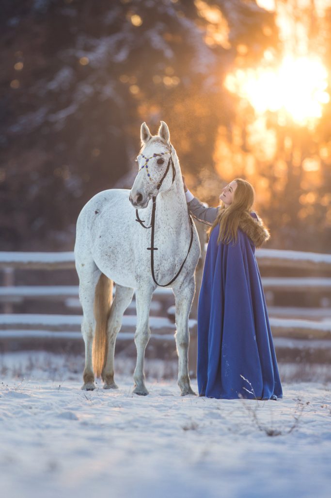 Eine Frau in einem blauen Mantel steht neben einem weißen Pferd im Schnee.