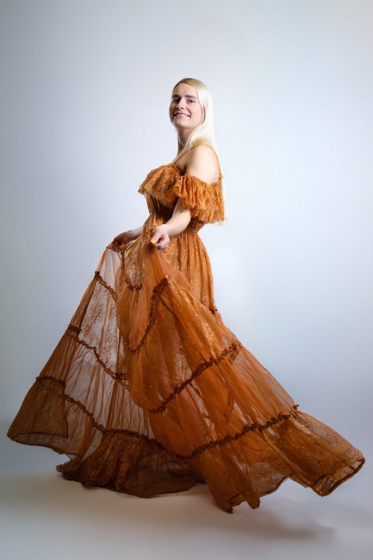 Frau in einem braunen, schulterfreien Kleid mit voluminösem Rock, vor neutralem Hintergrund.