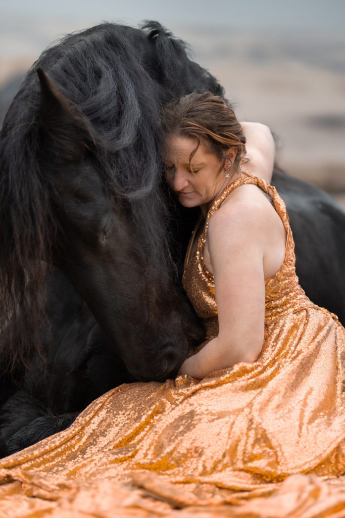 Eine Frau in einem orangefarbenen Kleid umarmt ein schwarzes Pferd am Strand.