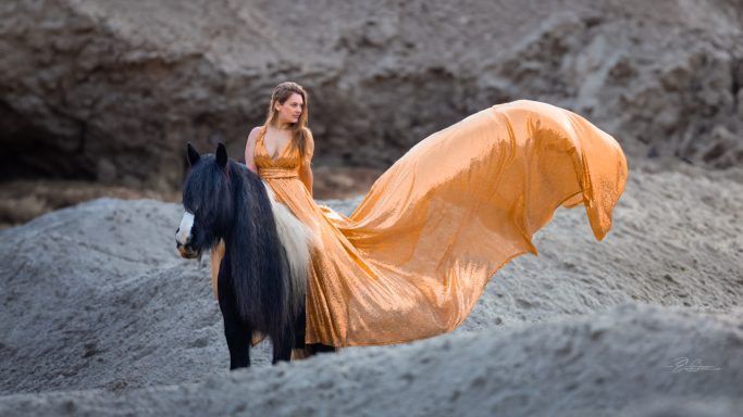 Frau in orangeem Kleid steht neben einem schwarzen Pferd in einer sandigen Umgebung.