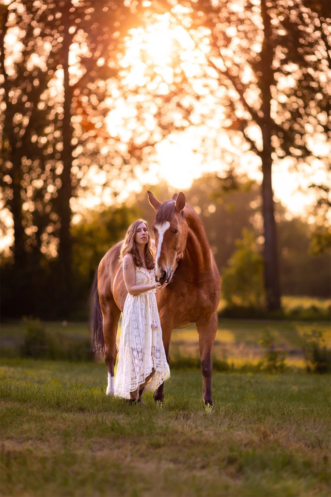 Ein Mädchen in einem weißen Kleid steht neben einem Pferd bei Sonnenuntergang.
