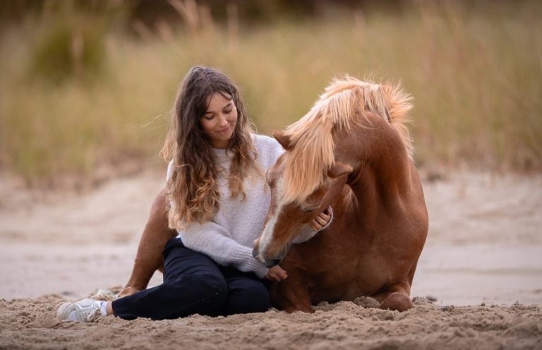 Junge Frau sitzt am Strand und kuschelt mit einem Pferd.
