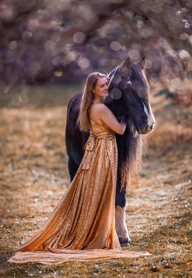 Frau in goldenem Kleid umarmt schwarzes Pferd im Freien.